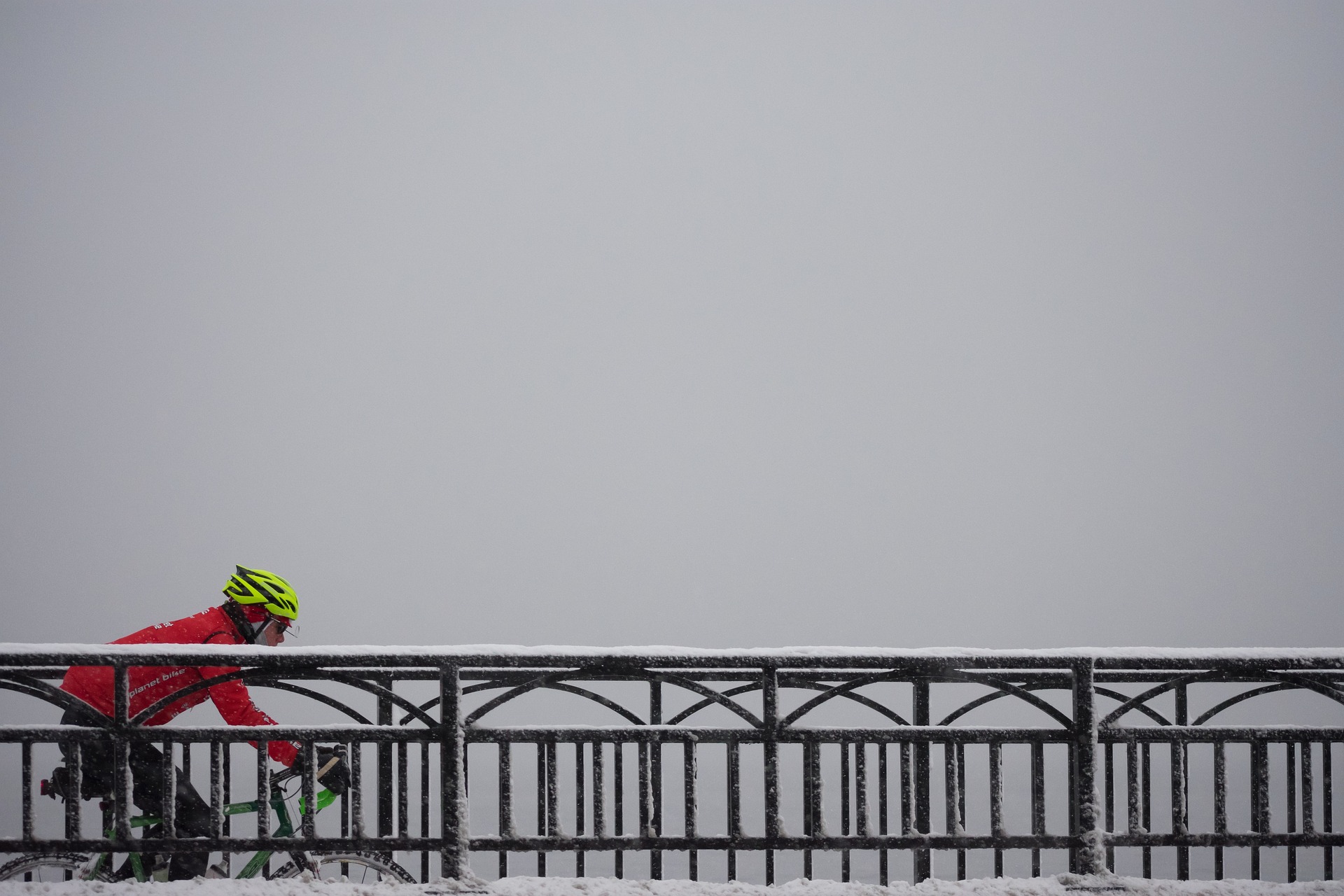 Rennradfahrer mit gelbem Helm und roter Jacke hinter einem schneebedeckten Brückengeländer. Im Hintergrund sieht man nur dristen Nebel